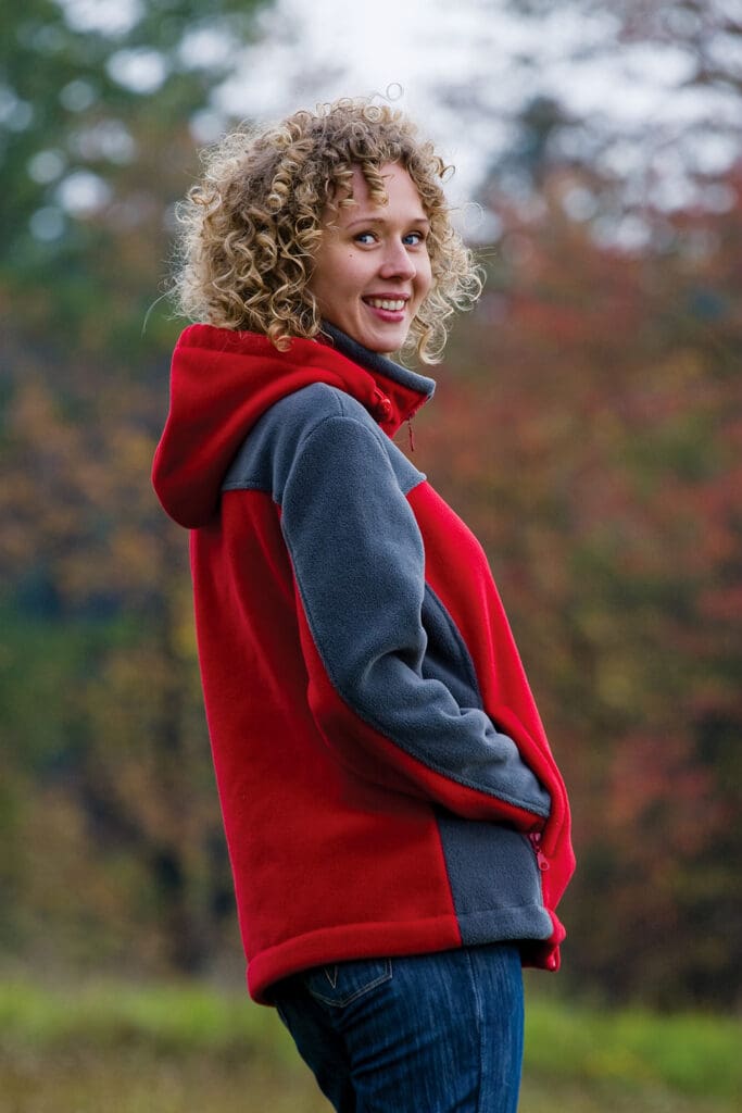 Modelka stojąca bokiem w polarze damskim fason VPL-10d, rozpinany na całości na zamek, stójka, dopinany kaptur, dwie kieszenie po bokach zapinane na zamek i z patką, kontrastowe wstawki modelujące sylwetkę, przykładowy kolor polaru czerwono-szary.